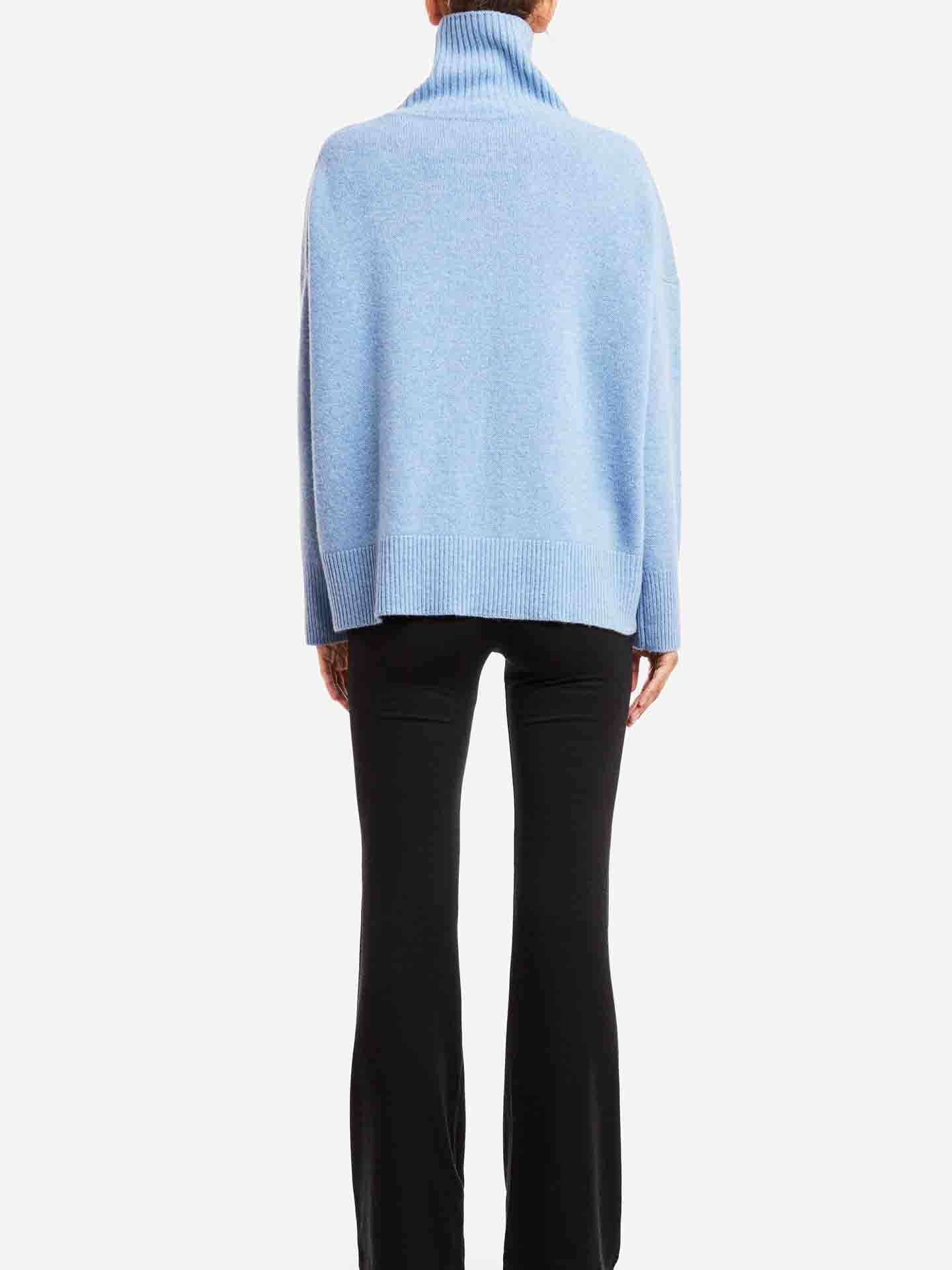 A person stands facing away from the camera, wearing a light blue Blefjell Sweater Women by We Norwegians and black pants. The luxurious piece appears soft and loose-fitting, hanging over the pants, which have a slight flare at the bottom. The background is plain white.