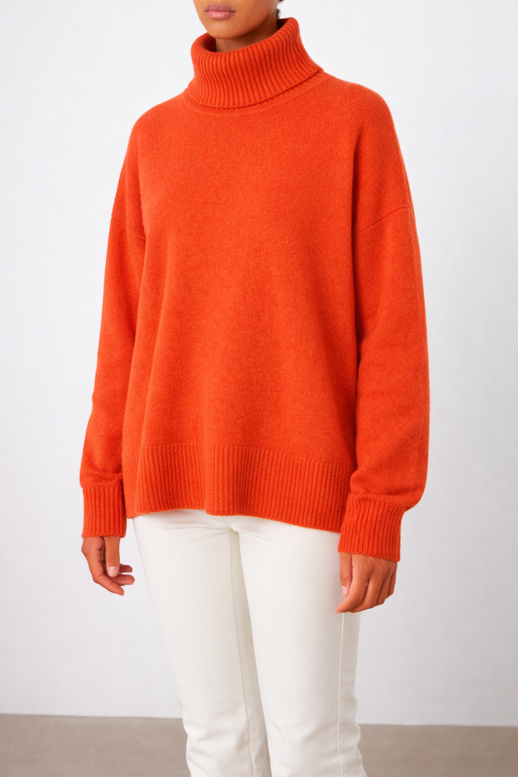 A woman models the We Norwegians Blefjell Sweater Women—Pure Cashmere in bright orange with long sleeves and ribbed cuffs, paired with white pants, against a plain light background—a modern luxury knitwear staple.