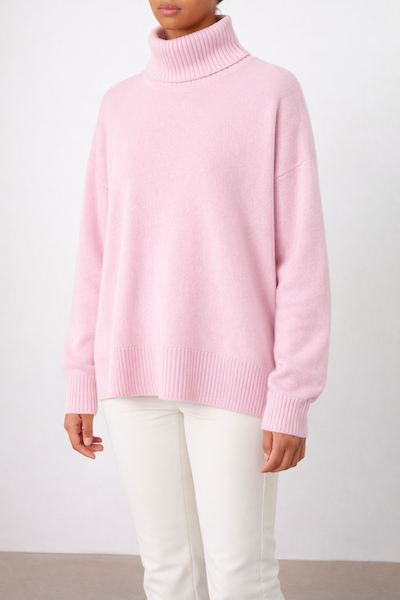 A person in the We Norwegians Blefjell Sweater Women - Pure Cashmere, a loose light pink top, and white pants stands against a plain light background with their face partially out of frame.