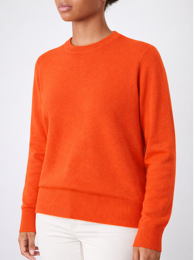 A person models the We Norwegians Blefjell Crewneck Women - Pure Cashmere in bright orange with white pants, standing against a plain light gray background. The image is cropped from the shoulders down, hiding the face.