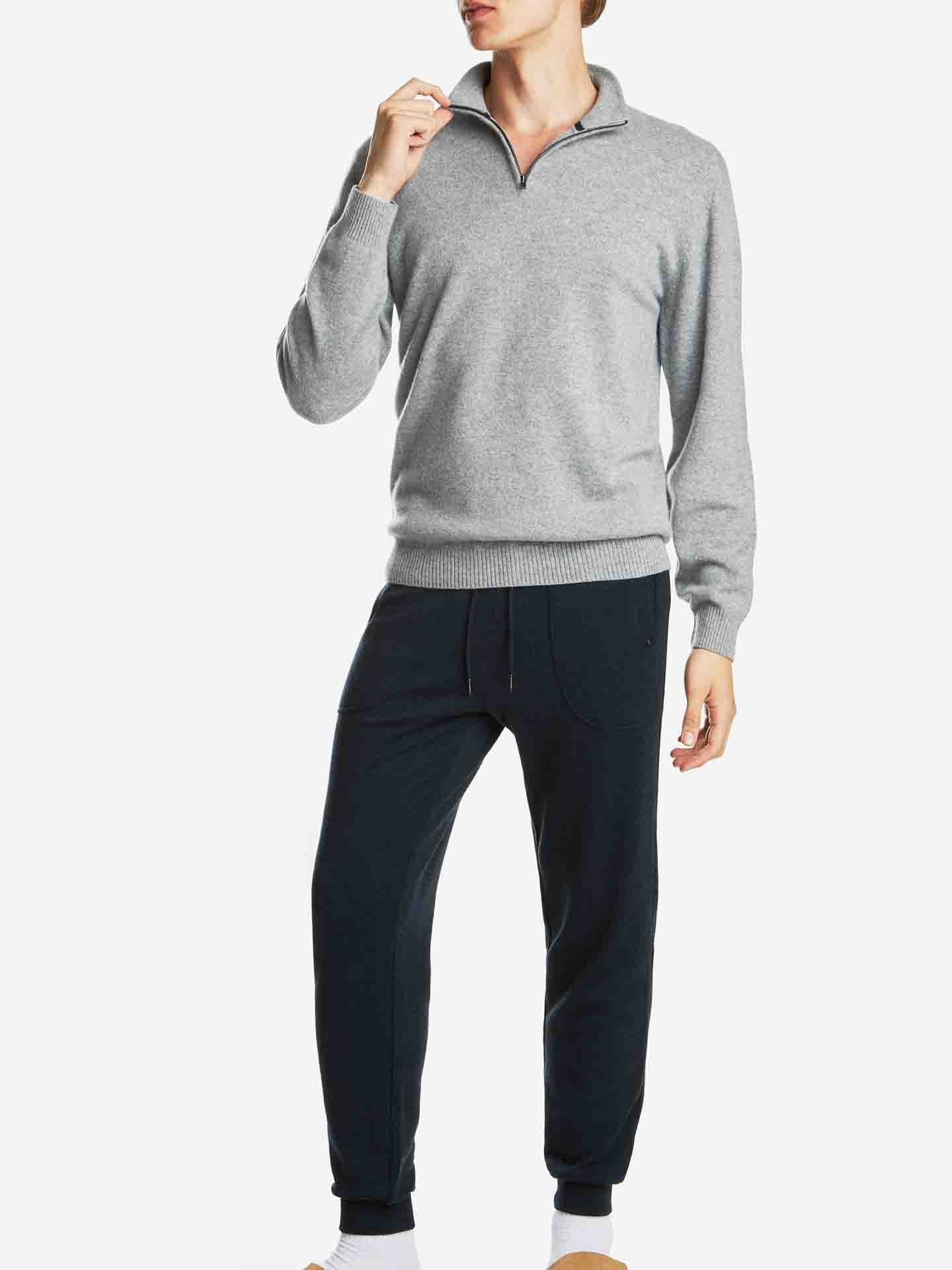 A person is posing against a plain white background, wearing the "Blefjell Zipup Men" light gray, long-sleeve cashmere sweater with a half-zip YKK zipper from We Norwegians and dark gray jogger pants. The person's left hand is raised to their chin, and they are wearing white shoes.