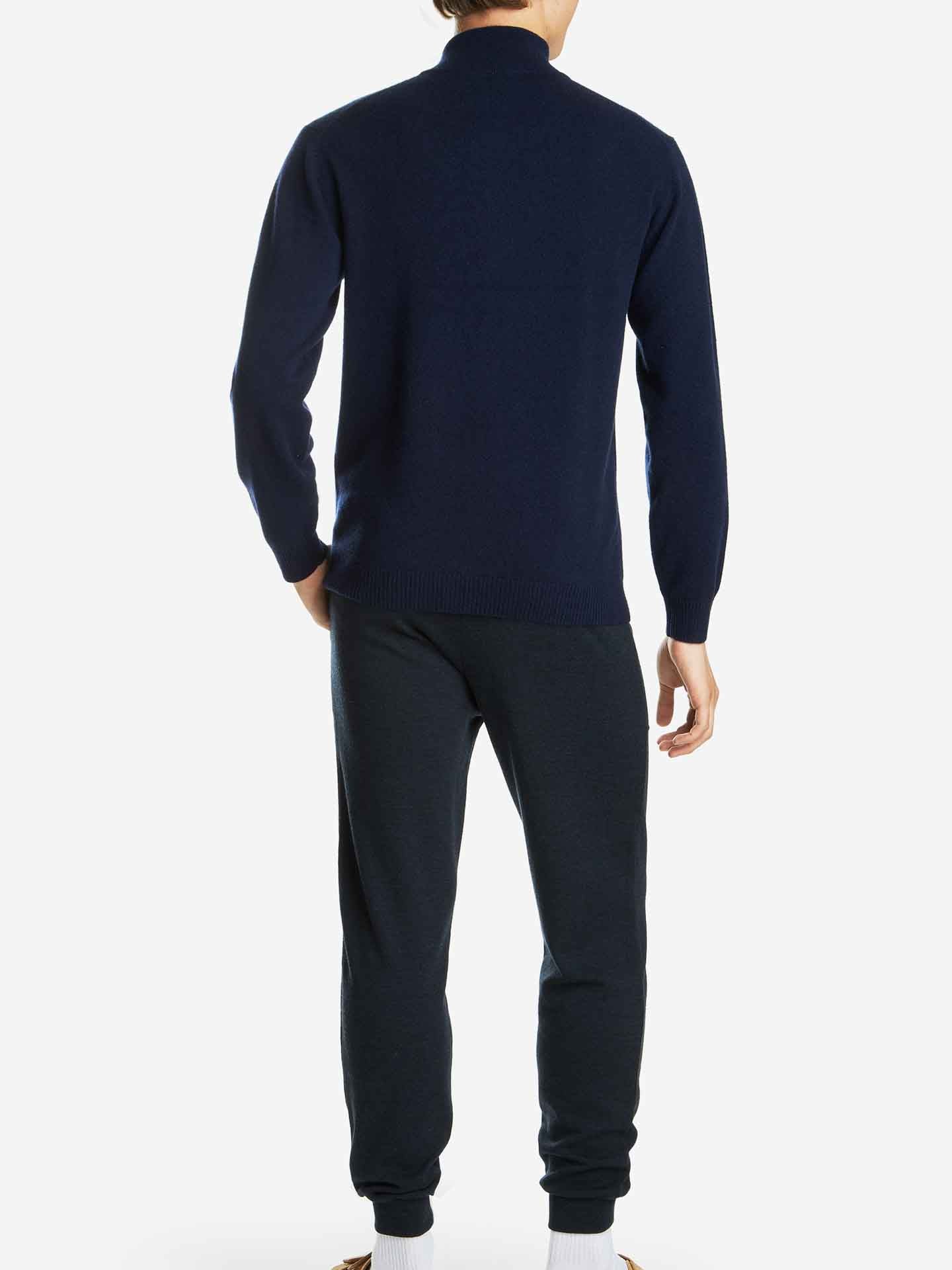 A person wearing the Blefjell Zipup Men navy blue long-sleeve shirt and matching navy blue pants from We Norwegians is standing with their back to the camera. The individual, showcasing a luxury staple in their attire, also wears white socks and is positioned against a plain white background.