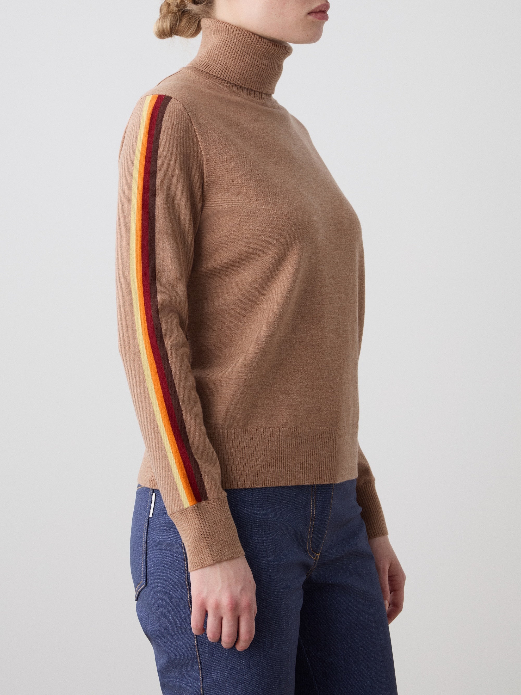 We Norwegians | FW25 | Womens Freebird Turtleneck Sweater_Cinnamon | Model is wearing this knitted turtleneck sweater with contrasting panels on the sleeves - paired with the Western ski pants | Shot against an off-white background | Side/ Close-up