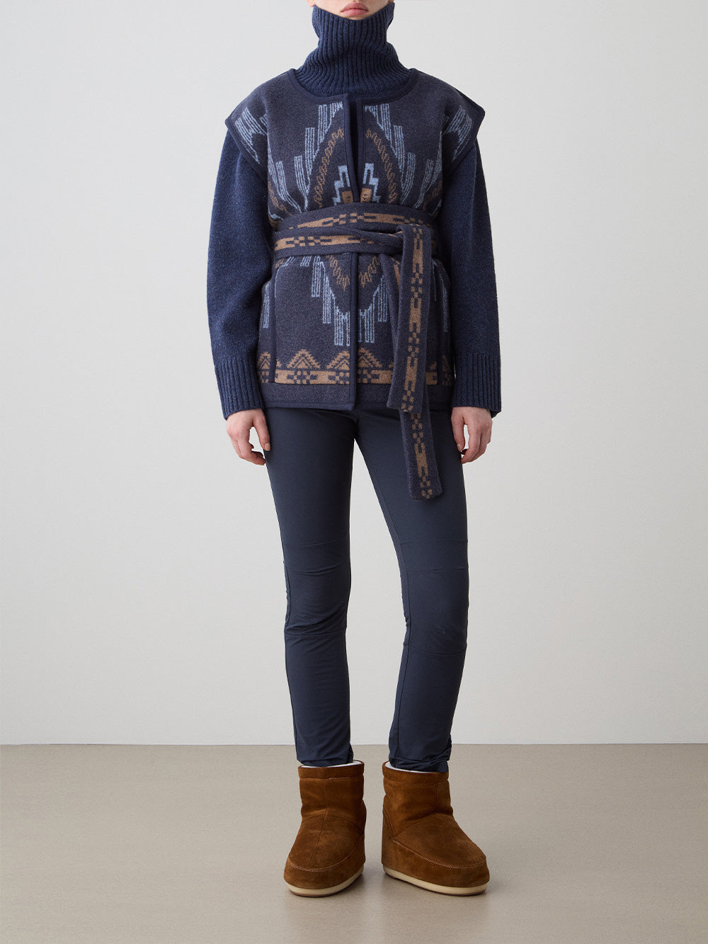 We Norwegians_FW25_Womens | Model is wearing the Kautokeino vest | Navy blue in color with a jacquard pattern inspired by Native and Indigenous American Art | Vest has a matching belt to cinch the waist | Front view on a white background