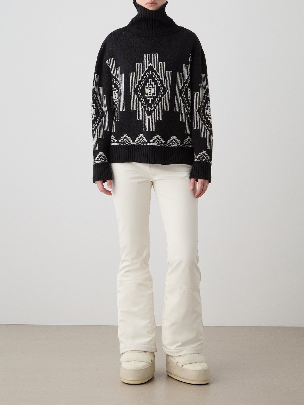 A woman wears the We Norwegians Kautokeino Sweater‚Äîblack wool with a white geometric pattern‚Äîpaired with off-white pants and boots, standing against a plain, light background.
