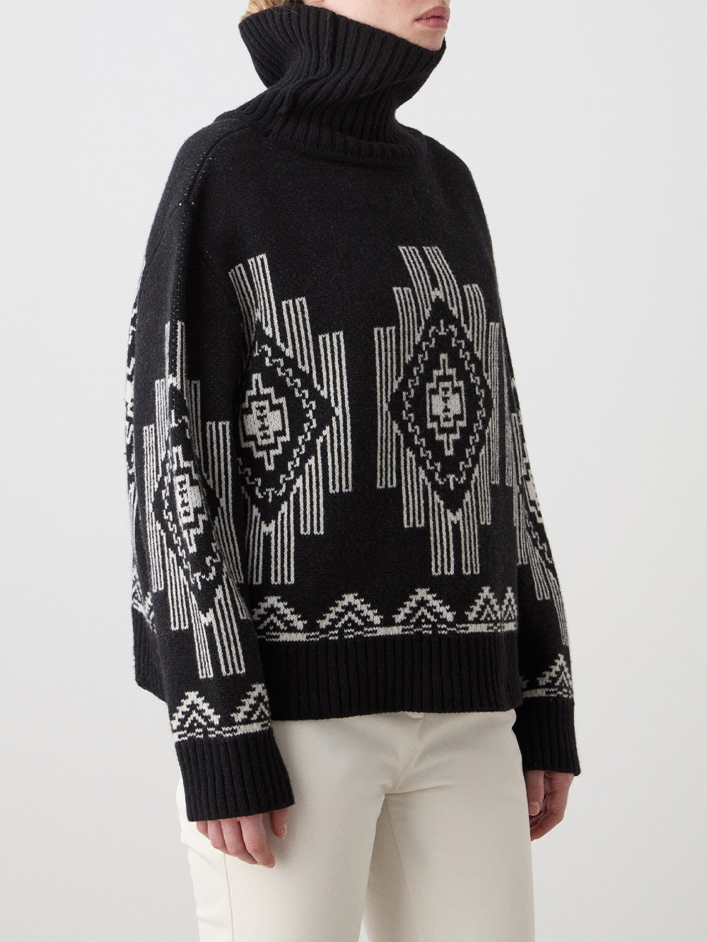 A woman wears the We Norwegians Kautokeino Sweater‚Äîa black wool apr√®s-ski knit with white geometric patterns, high collar, and long sleeves‚Äîpaired with cream-colored pants against a plain light background.