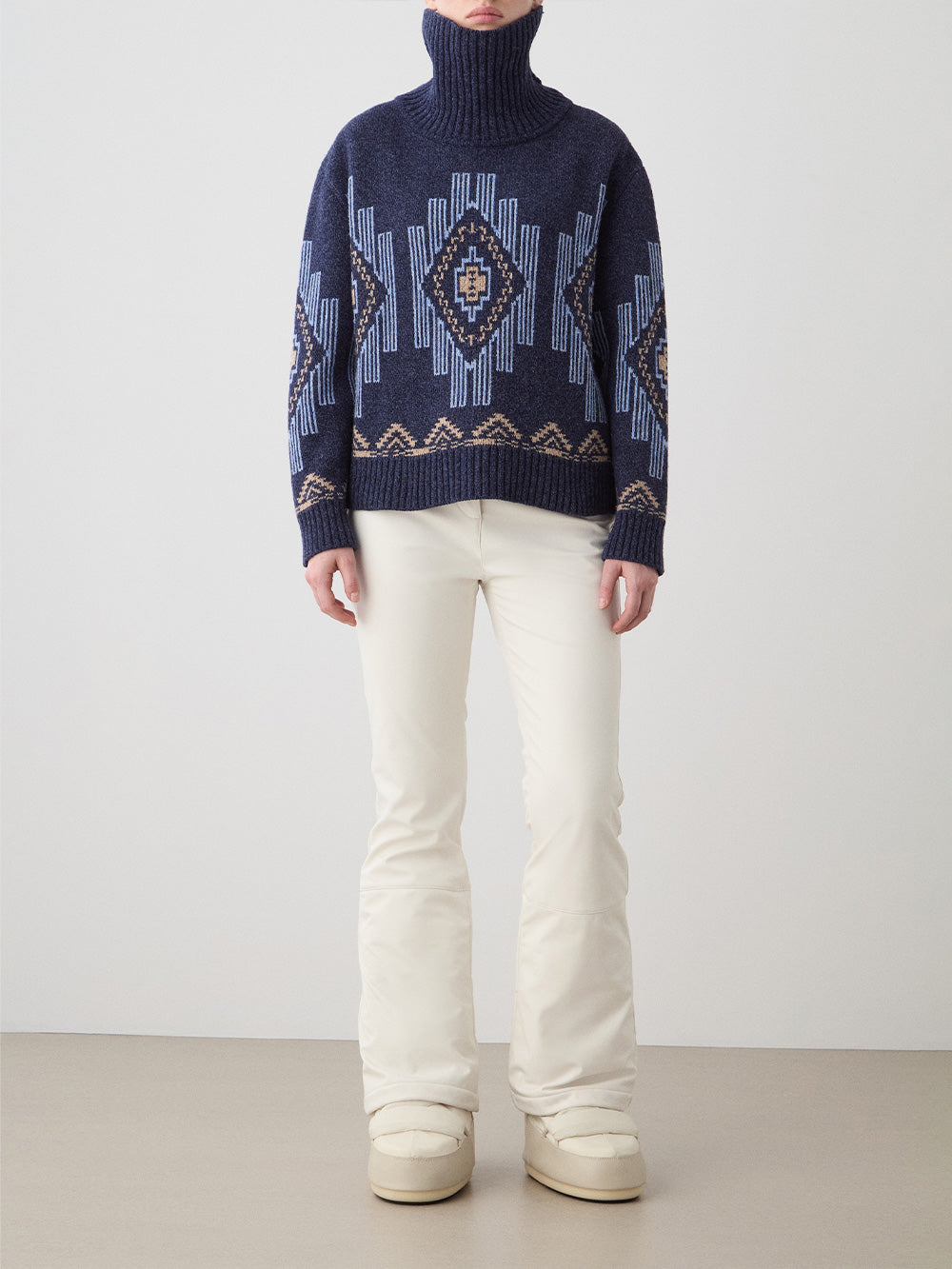 A person stands against a plain background wearing the We Norwegians Kautokeino Sweater women in navy with geometric patterns, paired with ivory pants and cream winter boots. Their face is not shown.