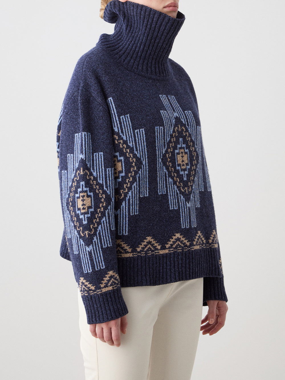 Model wearing the We Norwegians Kautokeino Sweater women in chunky navy blue with geometric light blue and tan patterns, paired with off-white pants, against a plain light background.