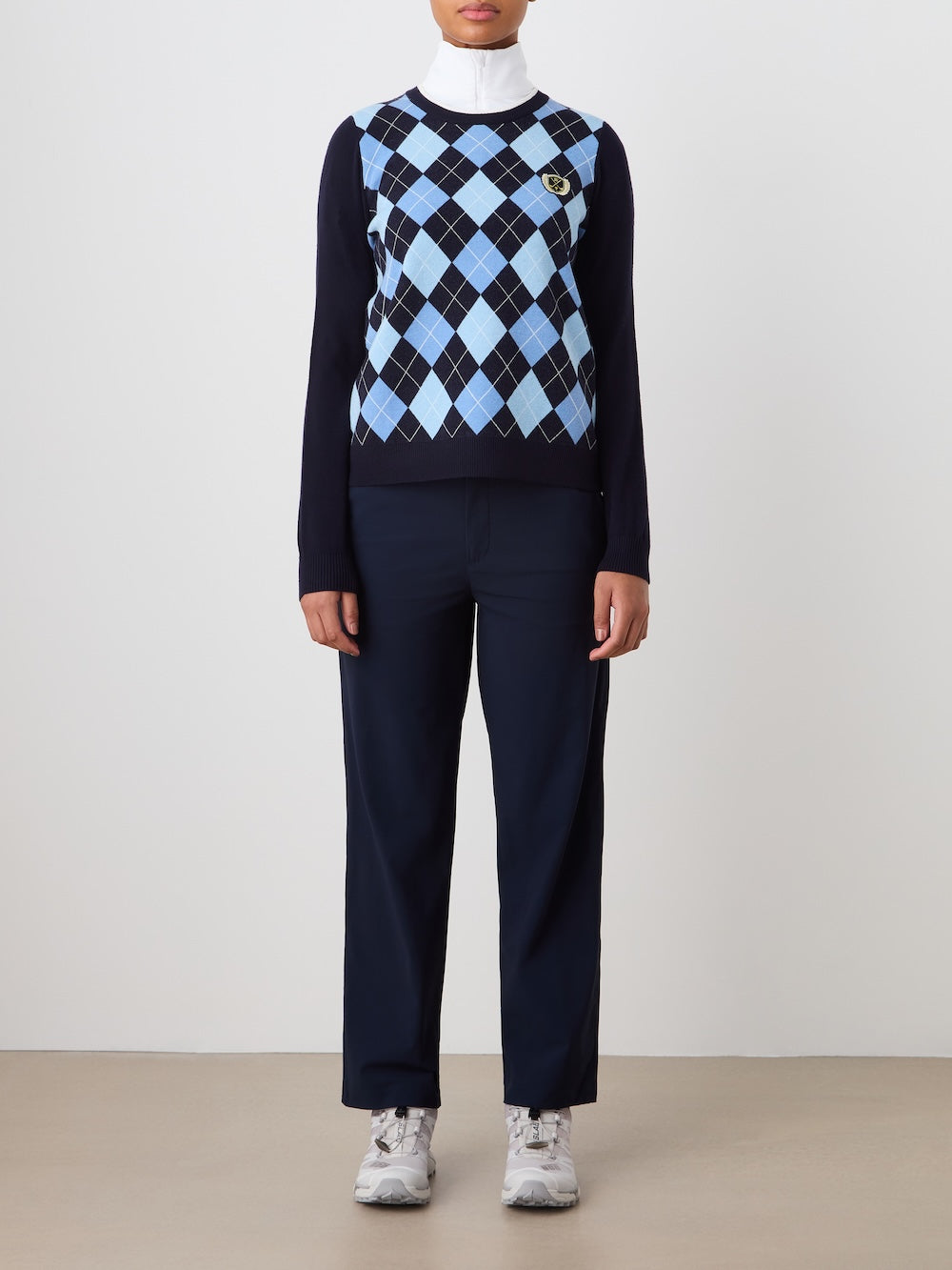 Person wearing the navy blue Fairway sweater with a blue argyle pattern and dark pants on a plain background_Front