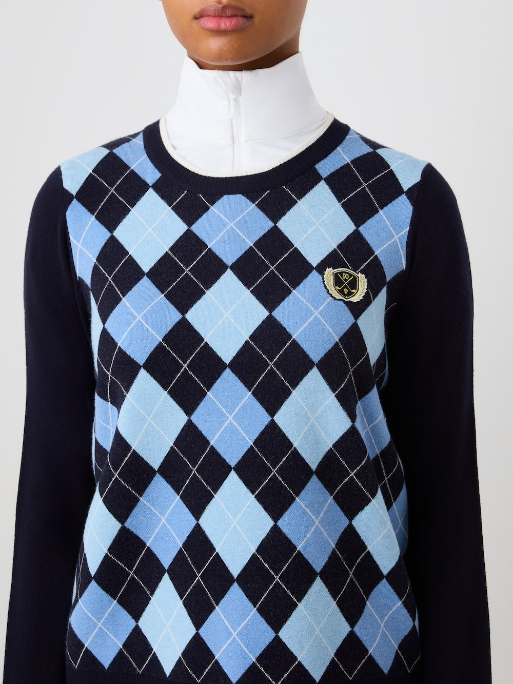 Person wearing the navy blue Fairway sweater with a blue argyle pattern over a white high neck top against a plain background.