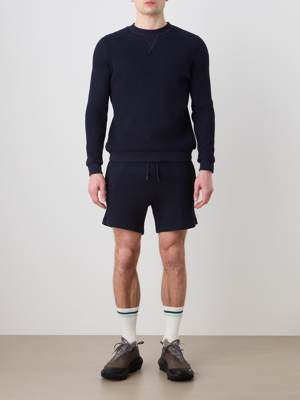 Person wearing a navy blue waffle crewneck and shorts with white socks and dark shoes against a plain background