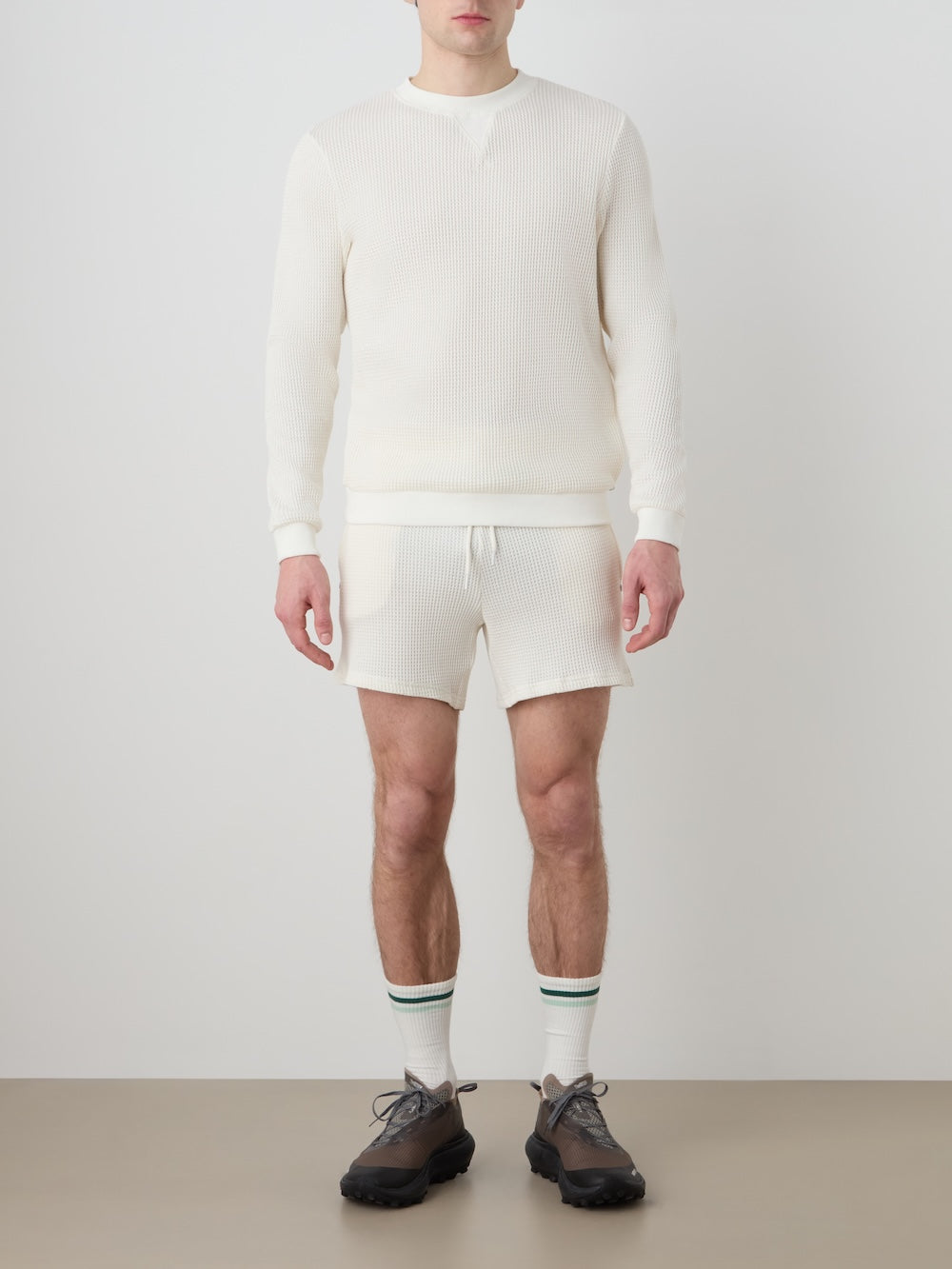 Person wearing an off white waffle crewneck with matching shorts, white socks and dark shoes on a light background