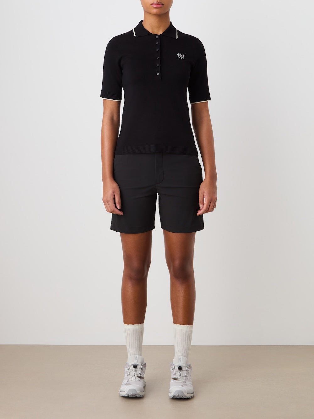 Person wearing a black polo shirt and shorts with white socks and shoes on a plain background