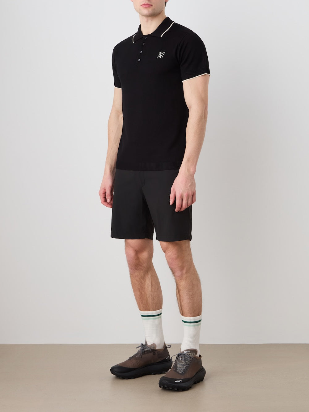 Person wearing a black polo shirt, black shorts, white socks with green stripes, and brown shoes on a plain background.